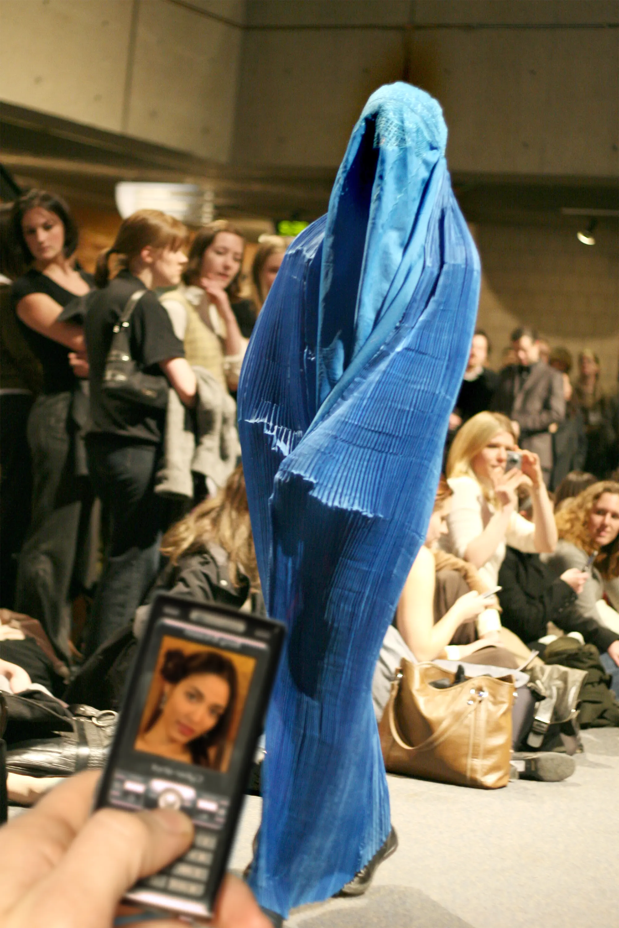 A lady in a burka during a catwalk. A mobile phone displays the face of the lady in the burka.